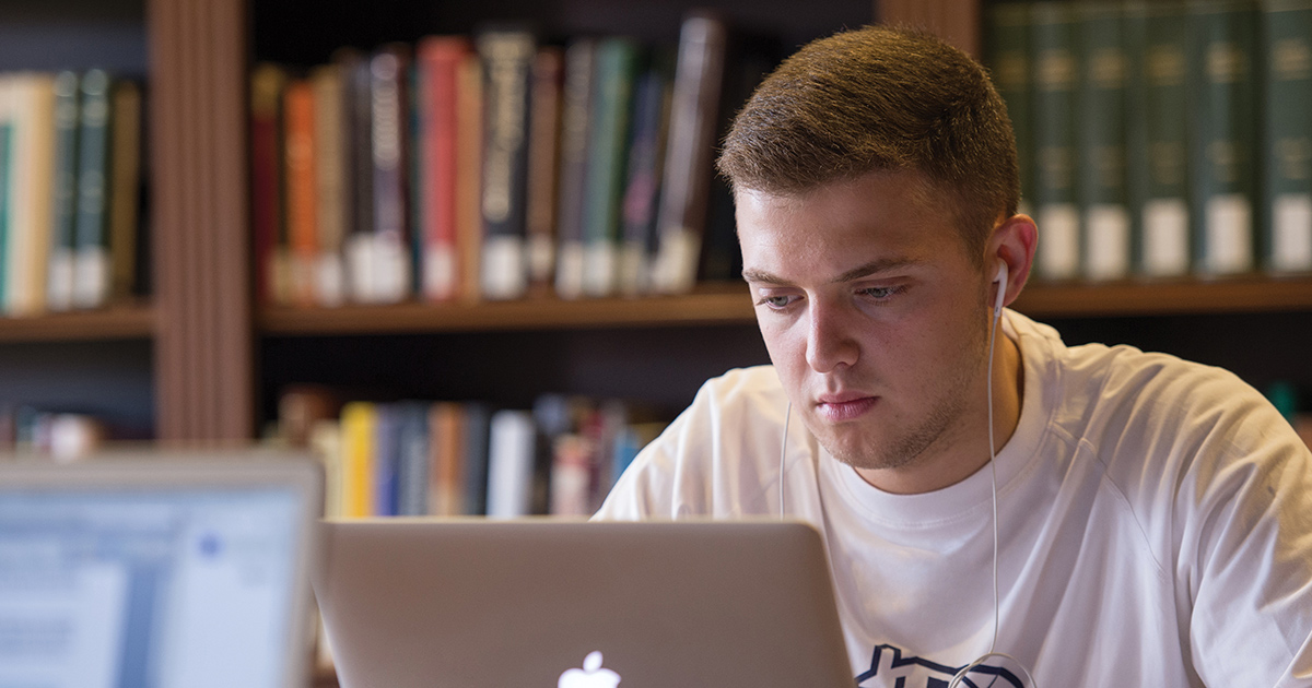 Student studying on a laptop while wearing white earbuds in front of colorful book shelf